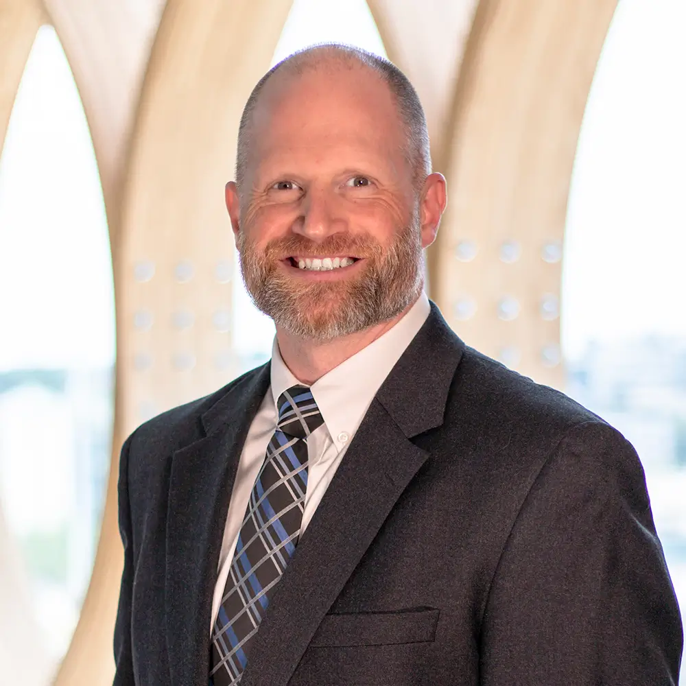 Smiling man with a beard wearing a dark suit, white shirt, and plaid tie, standing indoors in front of a bright, patterned background.