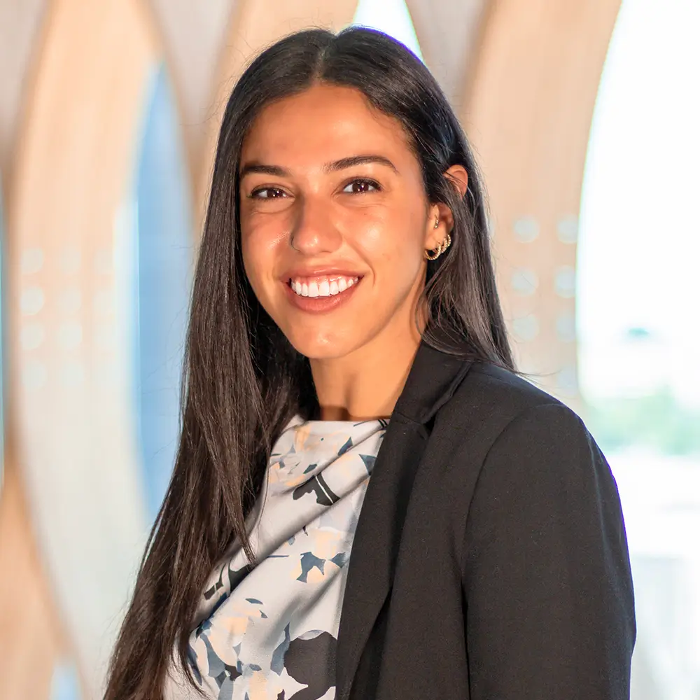 A woman with long dark hair, wearing a patterned top and black blazer, smiles in front of a softly lit, modern background with curved architectural elements.