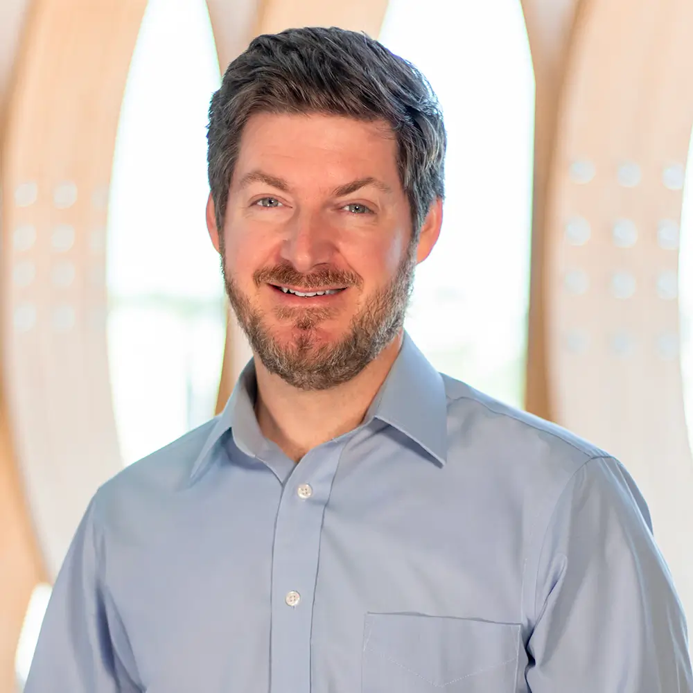 A man with short brown hair and a beard smiles while wearing a light blue button-up shirt. The background features soft, curved wooden shapes with bright natural light shining through.