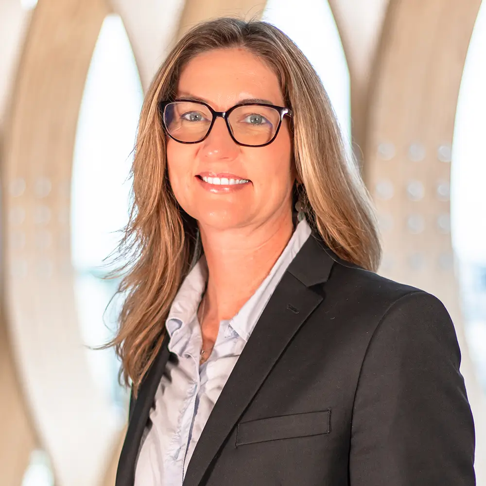 A woman with long brown hair and glasses wears a black blazer and light blouse, smiling in front of a bright, modern background with architectural details.