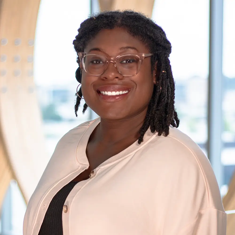A woman with glasses and shoulder-length twists smiles at the camera. She wears a light-colored jacket over a dark top. The background is softly blurred with large windows and natural light.